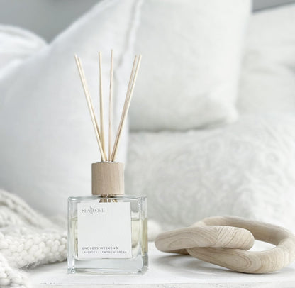 A serene and inviting space featuring an elegant reed diffuser with a label that says "endless weekend," placed beside a cozy white blanket and a wooden decorative object, creating an atmosphere of relaxation and tranquility.